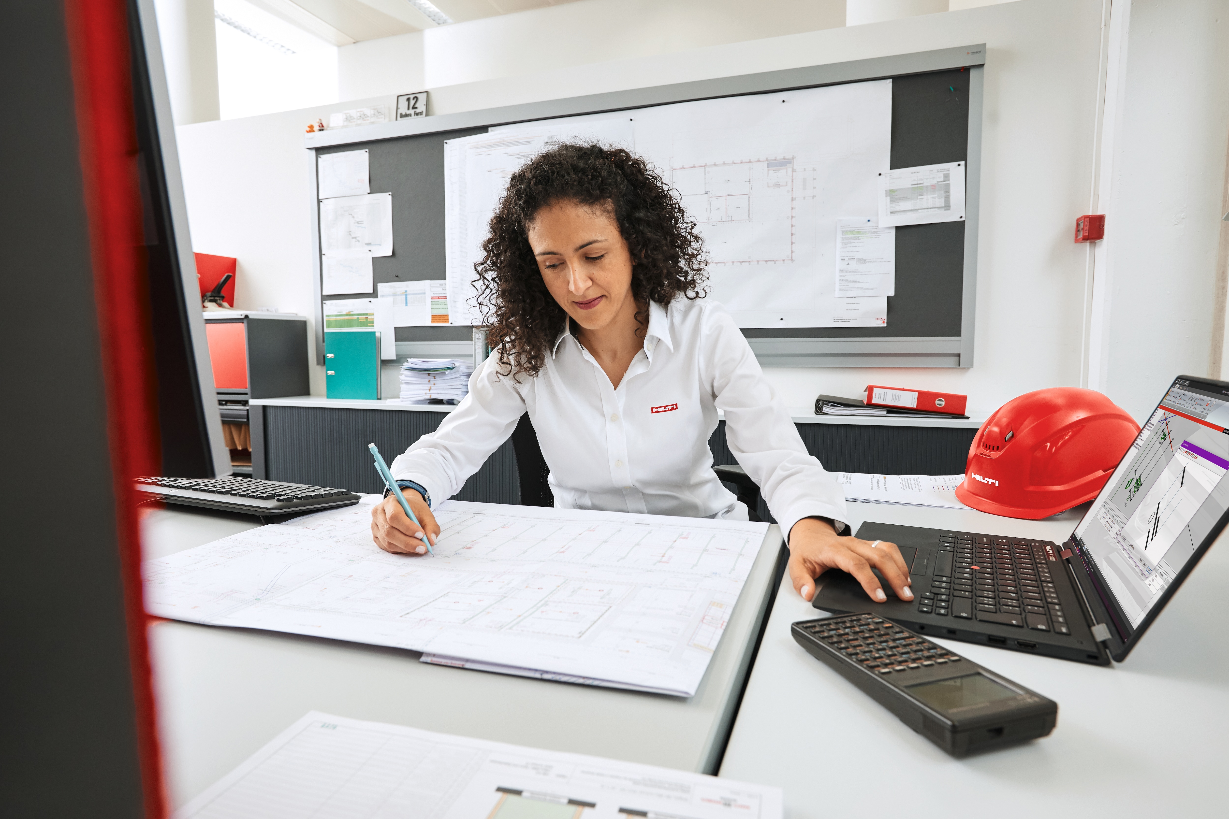 Woman working on laptop and paper plans in an office.
