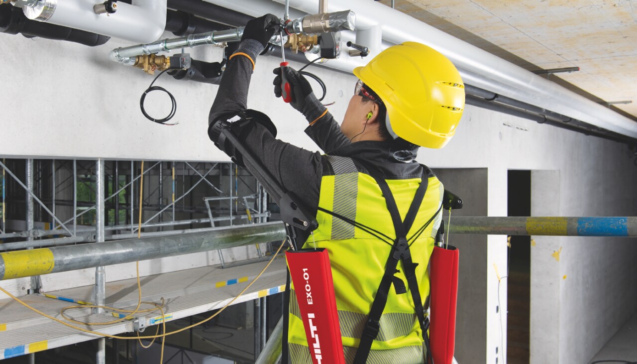Construction worker in yellow high visibility coat and helmet using Hilti EXO - T shoulder support and installing pipes into the ceiling.