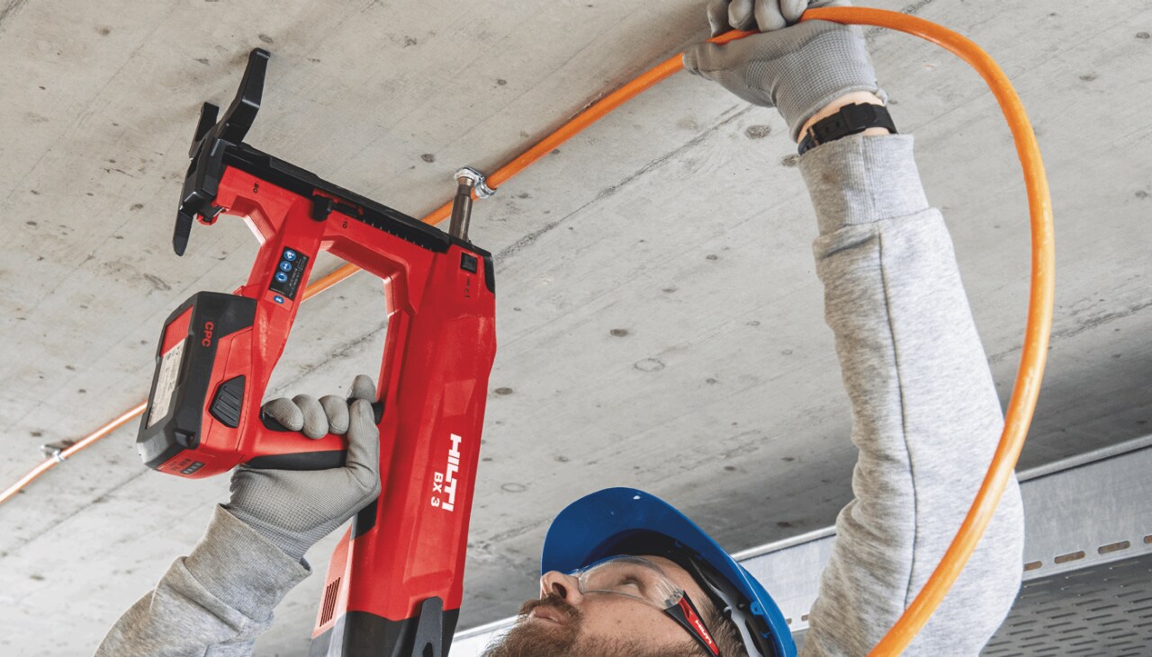 Close up of a man in blue helmet using red Hilti direct fastening to attached piping to ceiling.