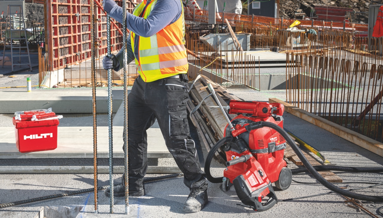 Construction worker in yellow high visibility vest installing rebar into concrete floor. Next to him are red Hilti power tools. 