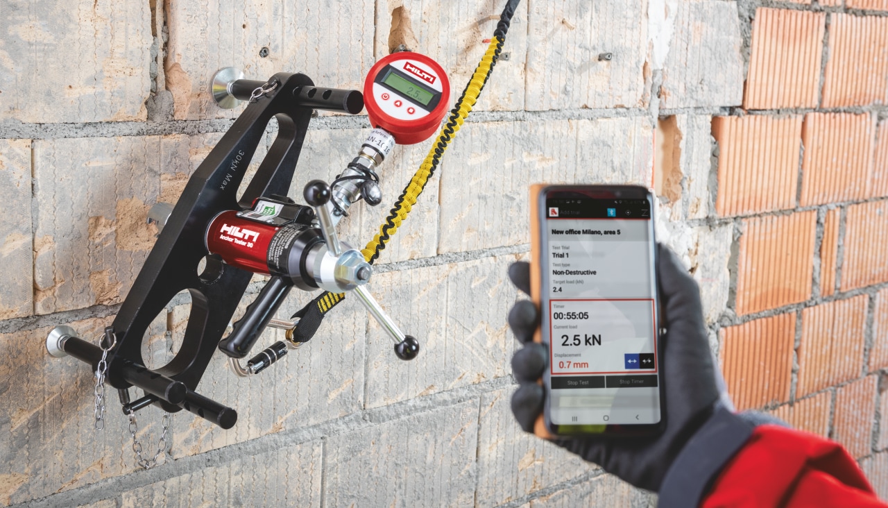 Close up of a hand holding smartphone with rebar resting app, next to it is a wall with rebar tester attached to rebar.