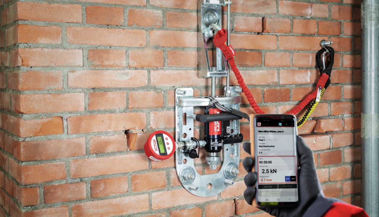 Close up of a hand holding smartphone with rebar resting app, next to it is a wall with rebar tester attached to rebar.