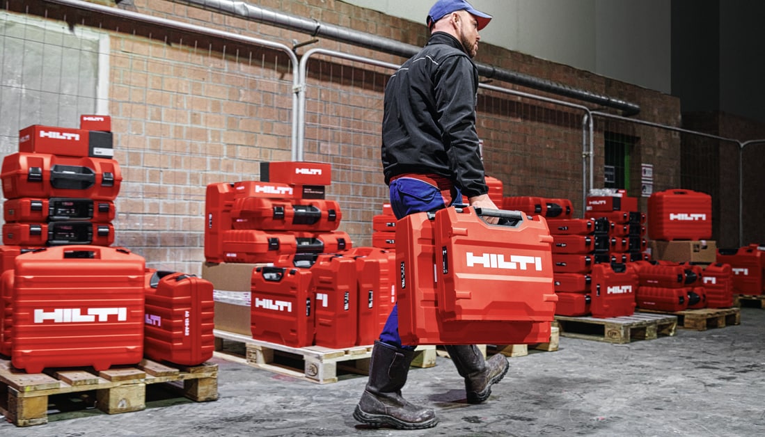 A PERSON CARRYING 2 HILTI TOOL CASES WITH MANY HILTI TOOLS ON PALLETS IN THE BACKGROUND OF A WAREHOUSE