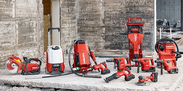 A variety of red and black power tools are neatly arranged on a concrete surface at a construction site. The tools include drills, a saw, and a vacuum, set against a backdrop of unfinished concrete walls.