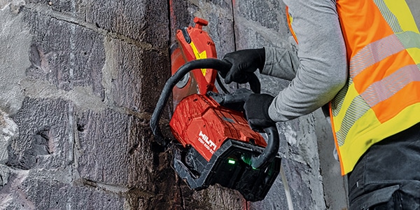 A construction worker wearing a high-visibility vest and gloves uses a red power saw to cut through a stone wall. The saw produces sparks as it cuts. The worker leans against the wall for support.