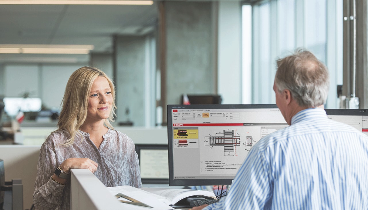 Woman talking to a man who is working in front of a computer screen