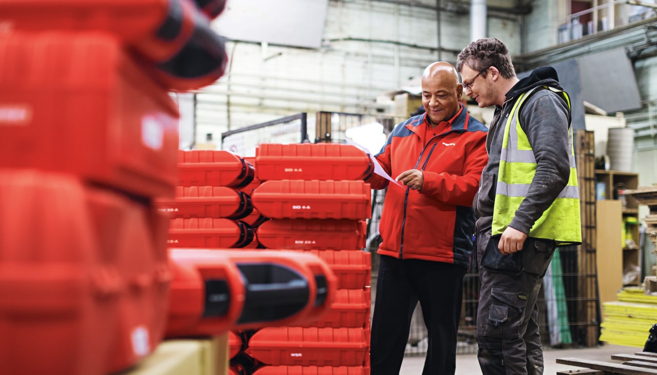 Hilti branded Account manager in red jacket talking to customer in high visibility vest. Next is many Hilti Red Tool boxes 