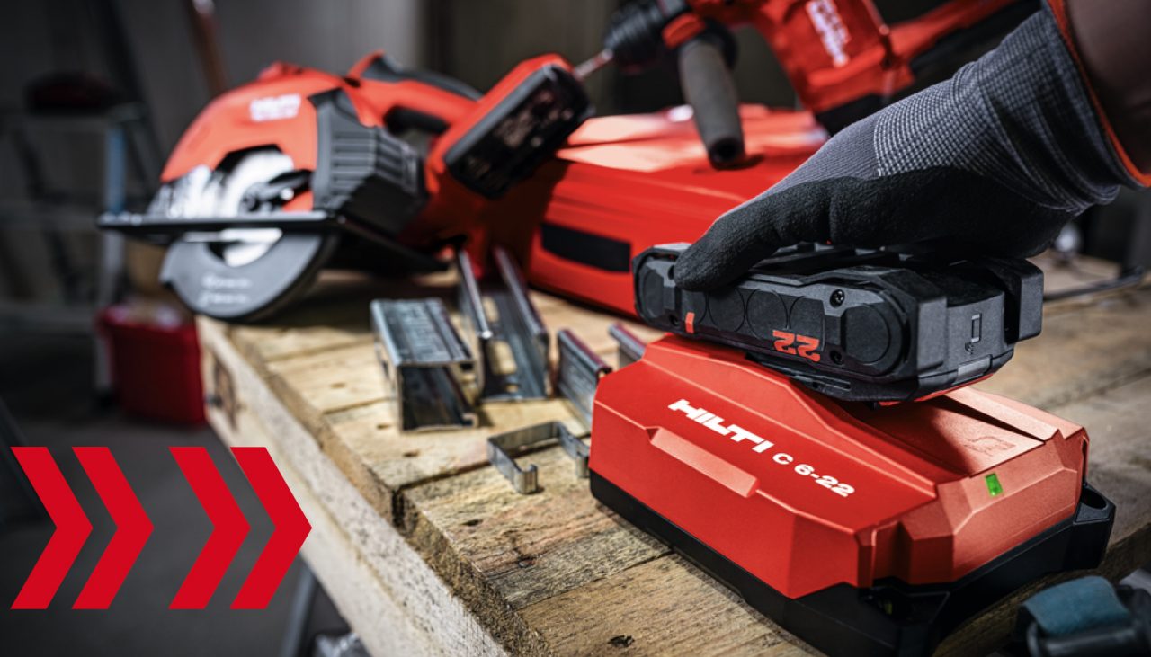 Construction table with Red Hilti saw, hammer drill and pieces of metal cuts. A man hand in safety glove is connecting Nuron battery to a charger.