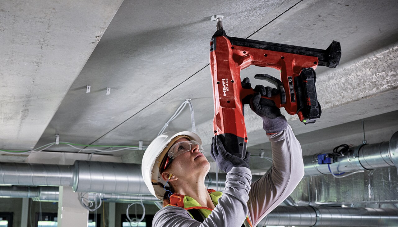 A woman in white construction helmet using red Hilti BX 4 -22 nail gun on the ceiling.