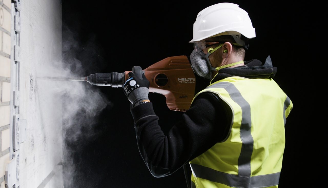 Man in yellow high visibility vest, white helmet and with respirator on moth is using red TE 700 AVR big demolition hammer on a wall.