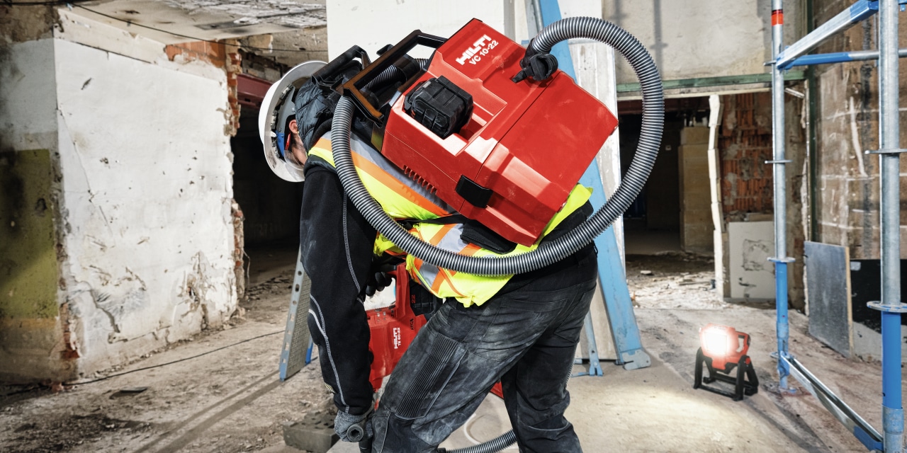 Construction worker in yellow high visibility vest having on his back, Hilti cordless vacuum cleaner as a backpack. 