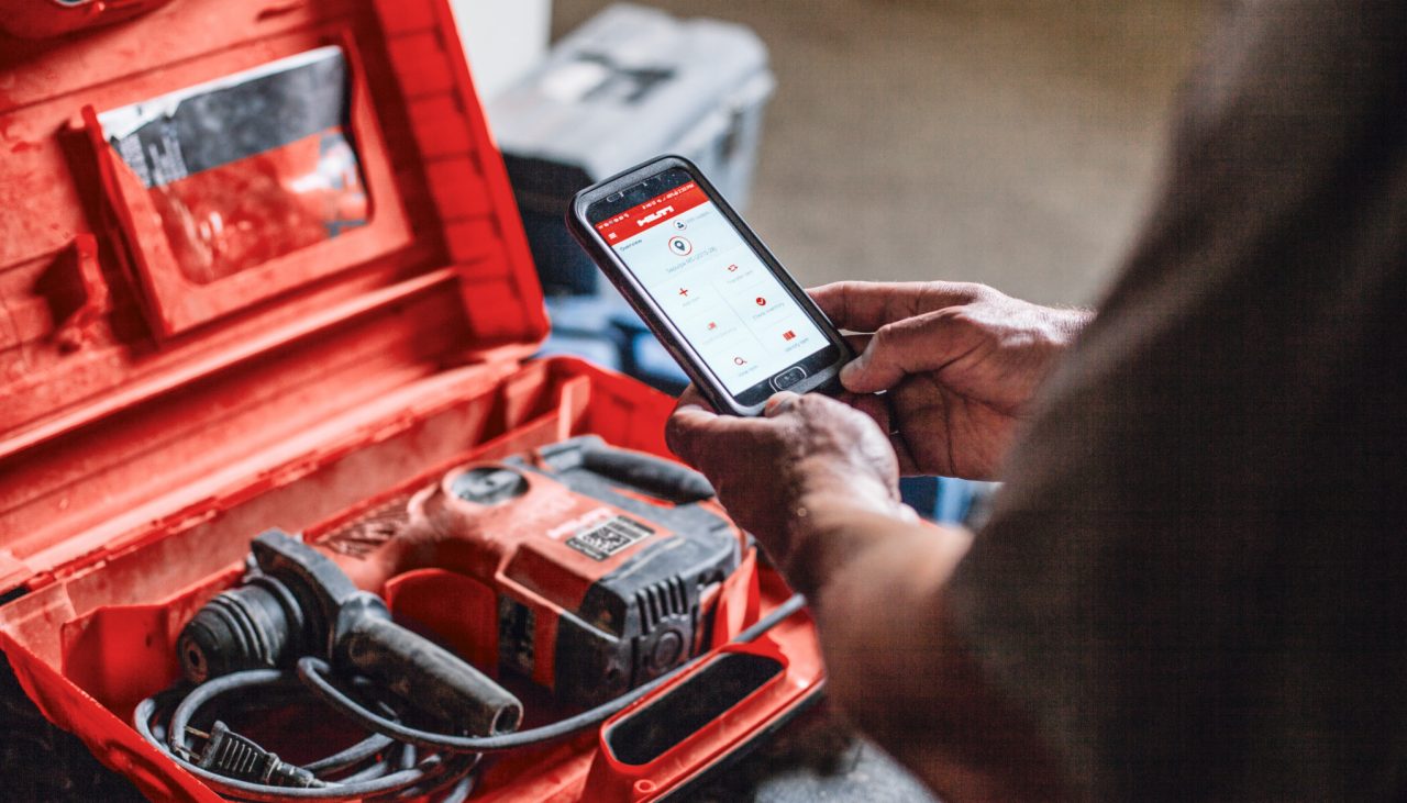 Man having smartphone with OnTrack app on the screen. Next to him is a Hilti red toolbox with a hammer drill in it.
