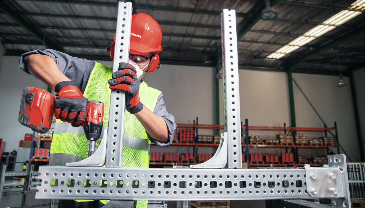 Construction worker in yellow high visibility vest using red Hilti screw driver and assembling metal support racking.