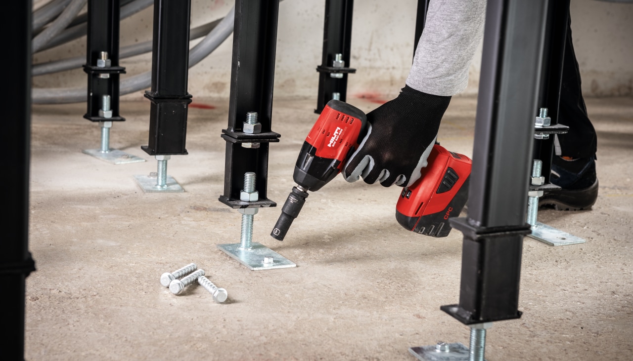 A worker installing large screws to concrete floor. The screws are holding black metal construction.