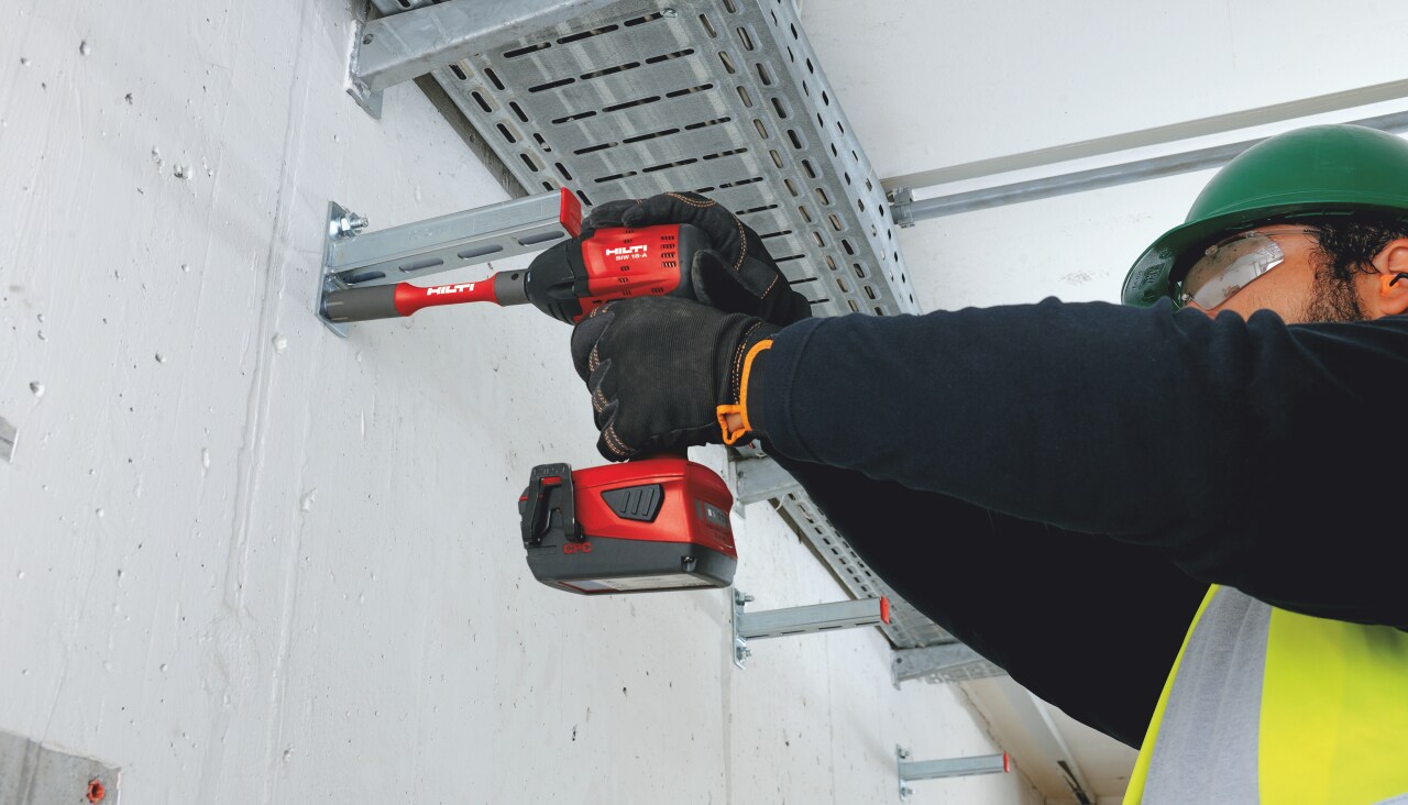 Construction worker in yellow high visibility vest and helmer is fixing metal construction to a concrete wall with hilti Red drill driver power tool.