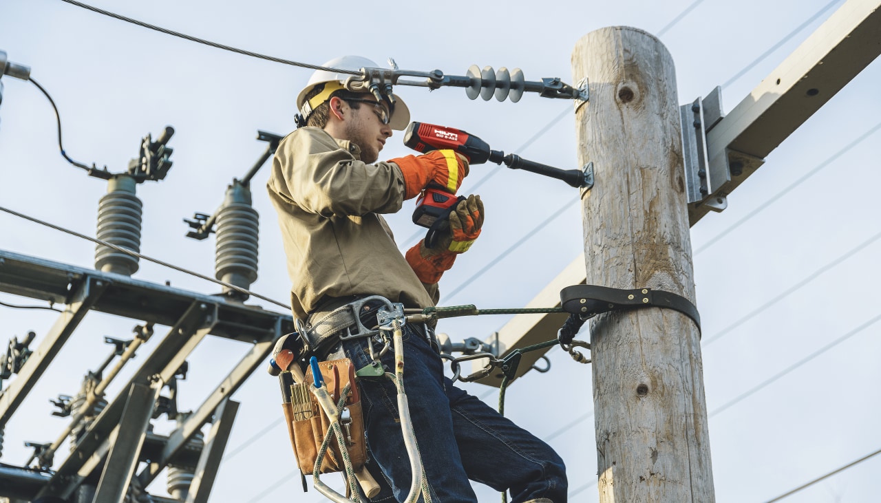 Construction worker in yellow high visibility coat and helmet using Hilti Red drill driver to drill a hole to a wooden construction holding power lines.