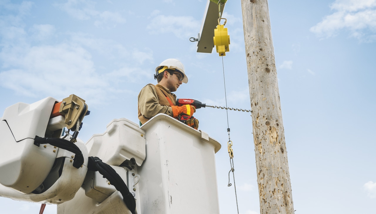 Construction worker in yellow high visibility coat and helmet using Hilti Red drill driver to drill a hole to a wooden construction holding power lines.
