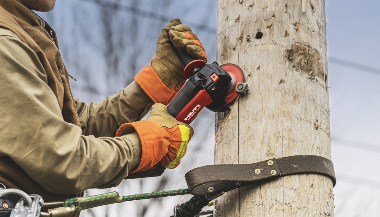 Construction worker in yellow high visibility coat and helmet using Hilti Red drill driver to drill a hole to a wooden construction holding power lines.Construction worker in yellow high visibility coat and helmet using Hilti Red angle driver to cut a metal screw from a wooden pilar.