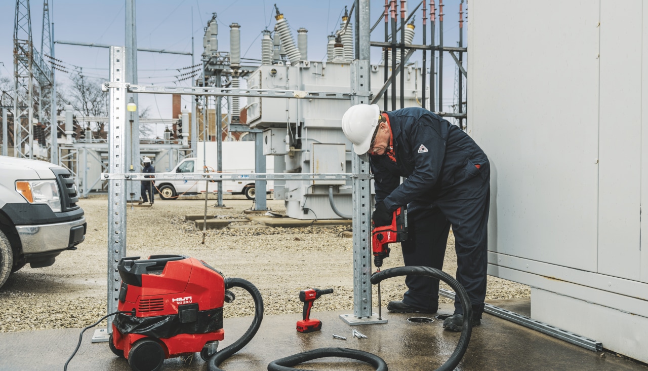 Construction worker is drilling a hole with Hilti red hammer drill into a concrete floor, in front of power plant.