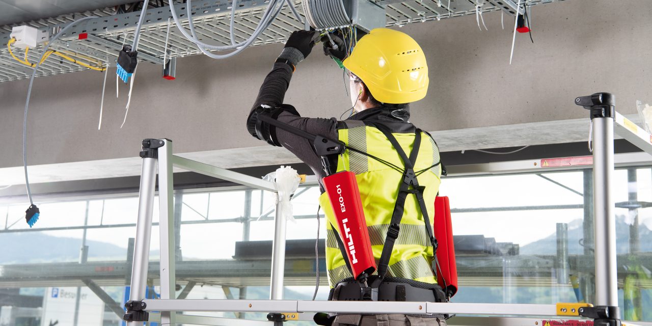 Man in yellow high visibility vest and helmet using red Hilti EXO-T shoulder support while installing cables to ceiling. 