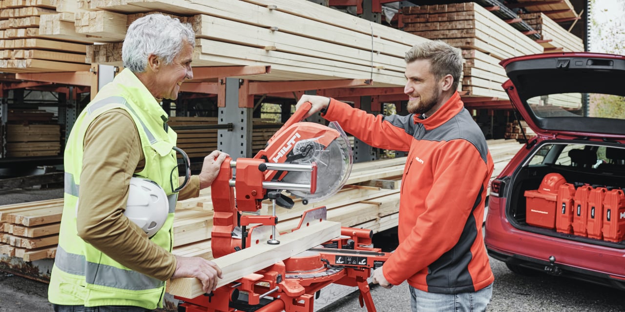 Two men discussing red Hilti Mitre saw in front of warehouse full of timber. On right hand side is red car with open back doors full of Hilti red tool boxes. 